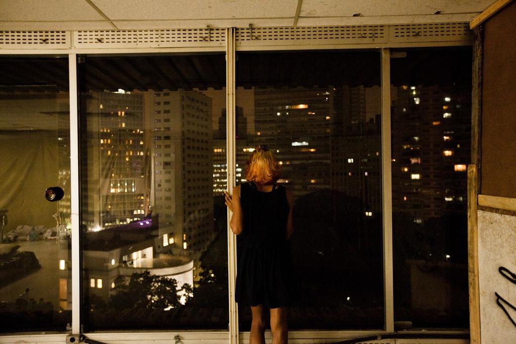 Uma travesti olha a cidade da janela do seu quarto.