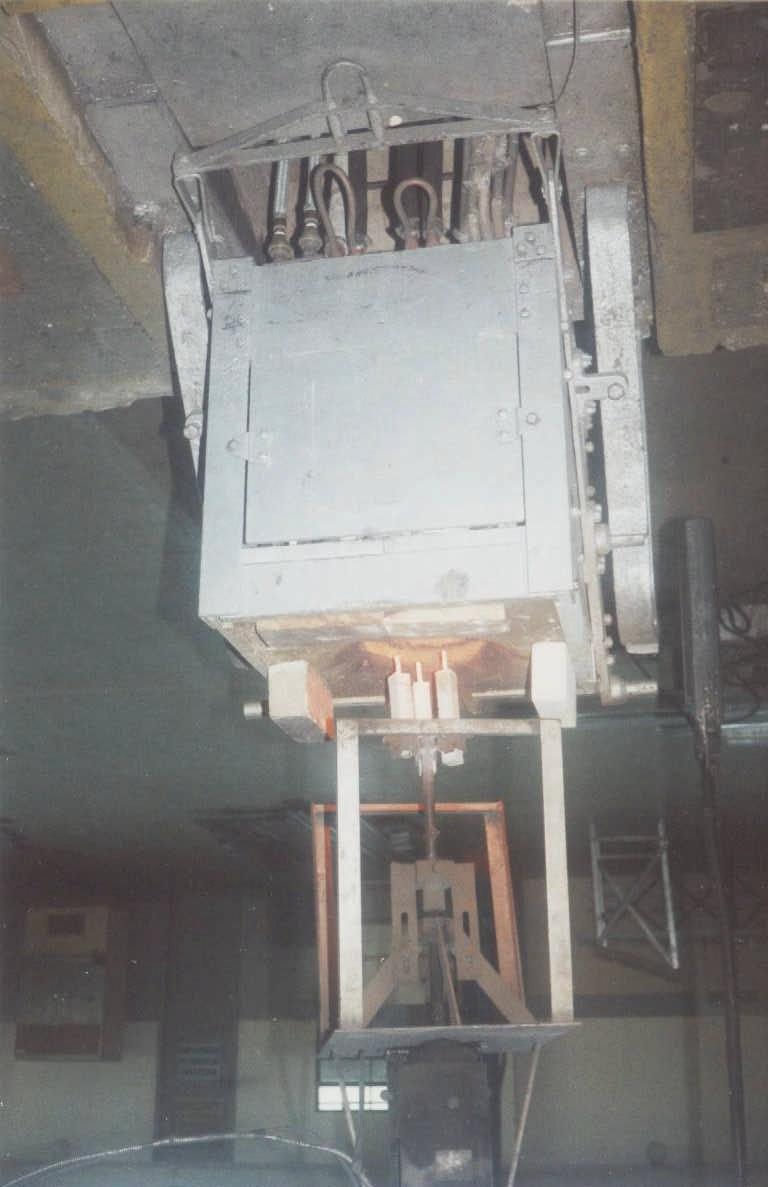 A figura 21 mostra uma vista do equipamento de teste, na qual se vê embaixo o forno de indução e acima, suspenso por uma ponte rolante, a estrutura onde se fixam as amostras e que possui um motor que