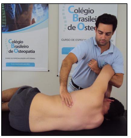 Osteopata em pé de frente para o paciente na altura da coluna torácica. A mão caudal toma contato (entre primeiro e segundo dedos) na costela inferior do espaço comprometido.