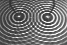 UNESP - 2019 A figura mostra um fenômeno ondulatório produzido em um dispositivo de demonstração chamado tanque de ondas, que neste caso são geradas por dois martelinhos que batem simultaneamente na